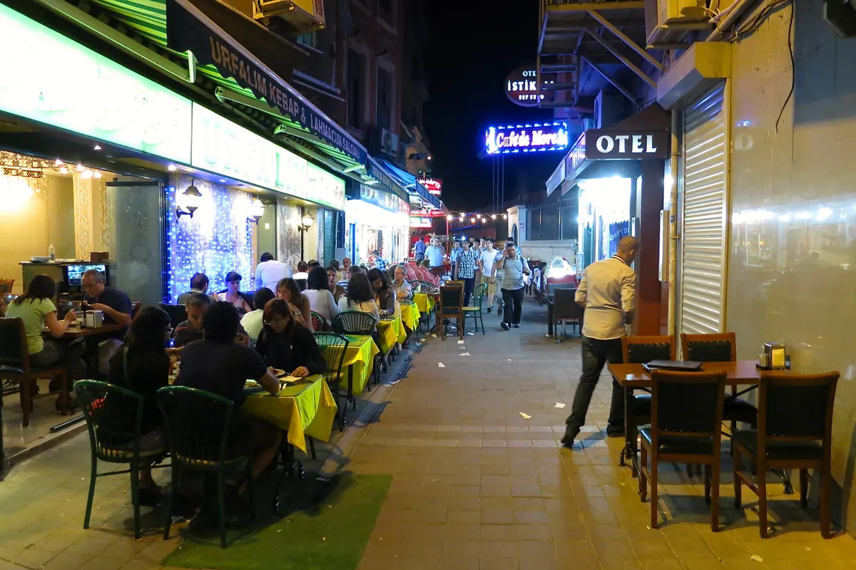 A pedestrian street in Istanbul at night, lined with restaurant tables under lights and signs, with diners and passers-by