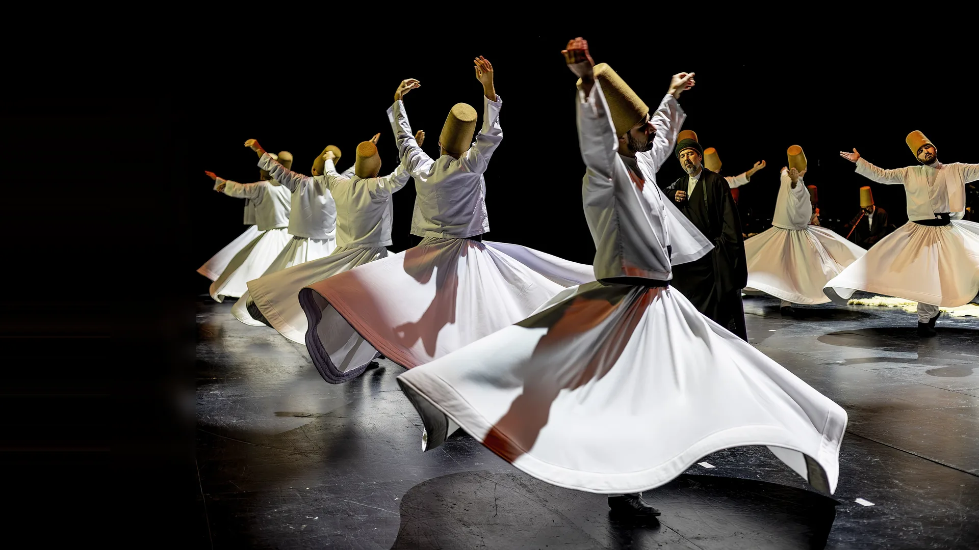 Semazens in long white skirts and tall brown felt hats whirling on a dark stage, arms raised, with a robed sheikh watching from the centre.